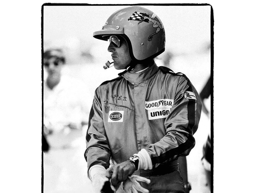 A black-and-white photograph of a race car driver wearing a helmet and racing suit. The helmet features a checkered flag and a cartoonish design on the side, and the suit is adorned with sponsor logos such as Goodyear, Union 76, and Hella. The driver's sunglasses and serious expression suggest intense focus, while he appears to be preparing for a race. The retro style of the equipment and attire hints at a historical motorsport era.